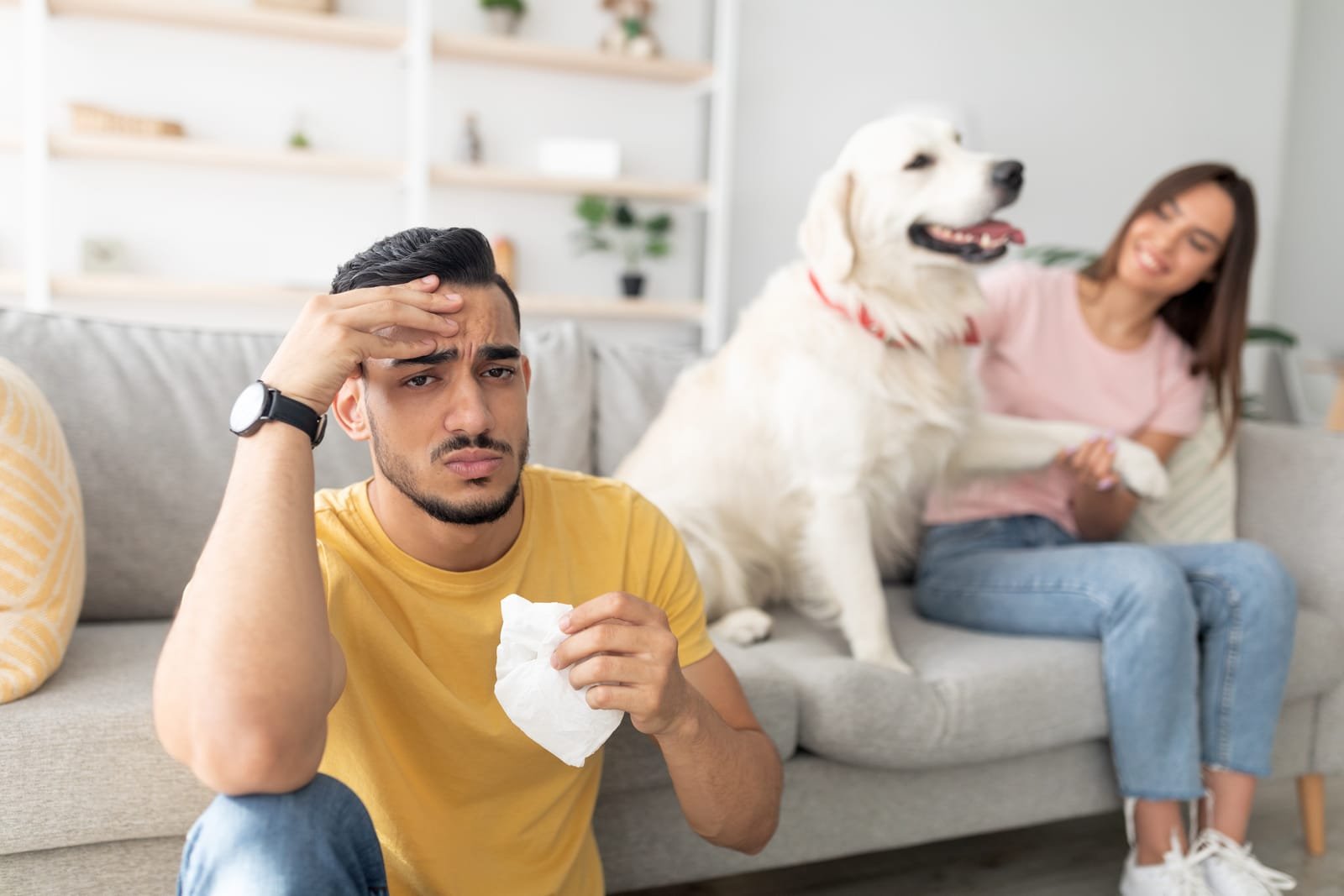 Young man looks worried while woman comforts him with a golden retriever nearby in a cozy living room setting